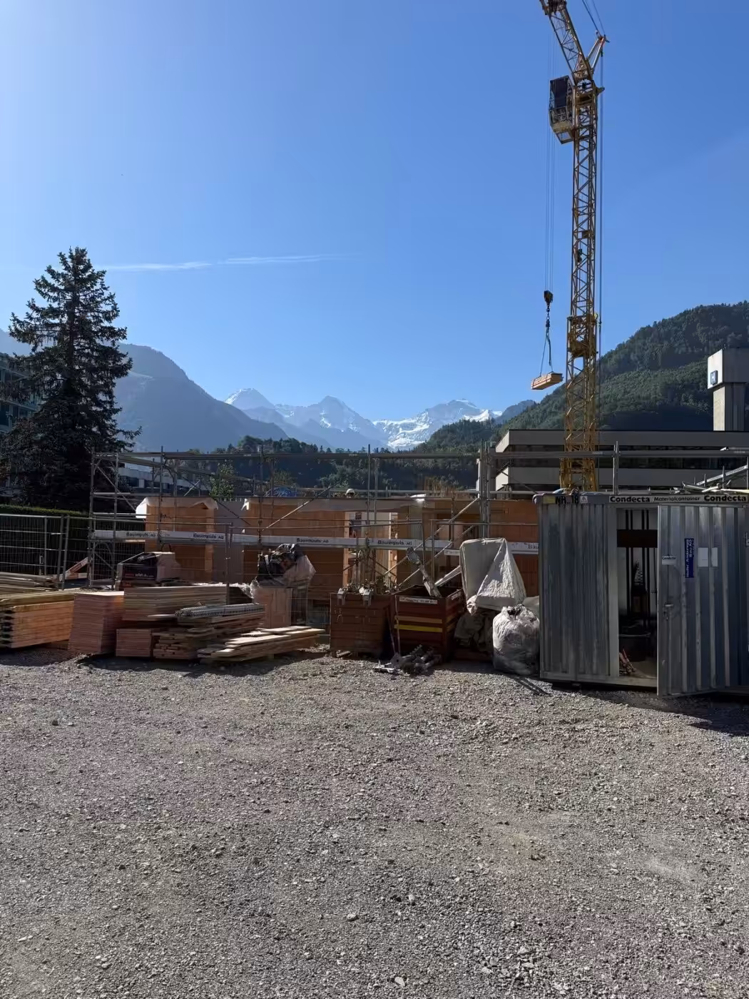 Baustelle mit Bergblick, Kran und Materialien unter klarem Himmel an einem sonnigen Tag.