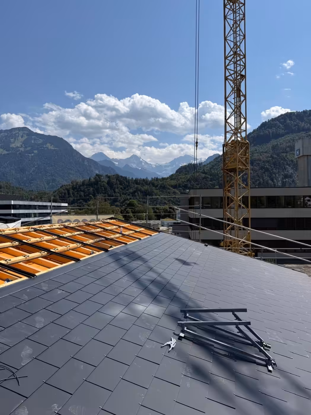 Dacharbeiten mit Kran im Bergpanorama, sonniger Himmel, modernes Gebäude in alpiner Landschaft im Hintergrund.