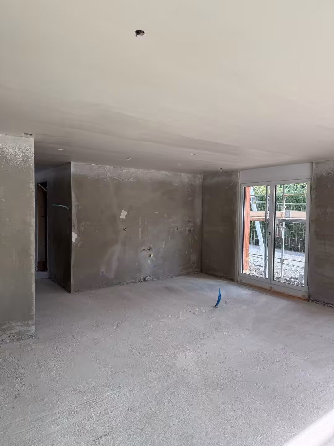Rohbau eines Zimmers mit verputzten Wänden, Fensterfront und unbehandeltem Betonboden, bereit für den Innenausbau.