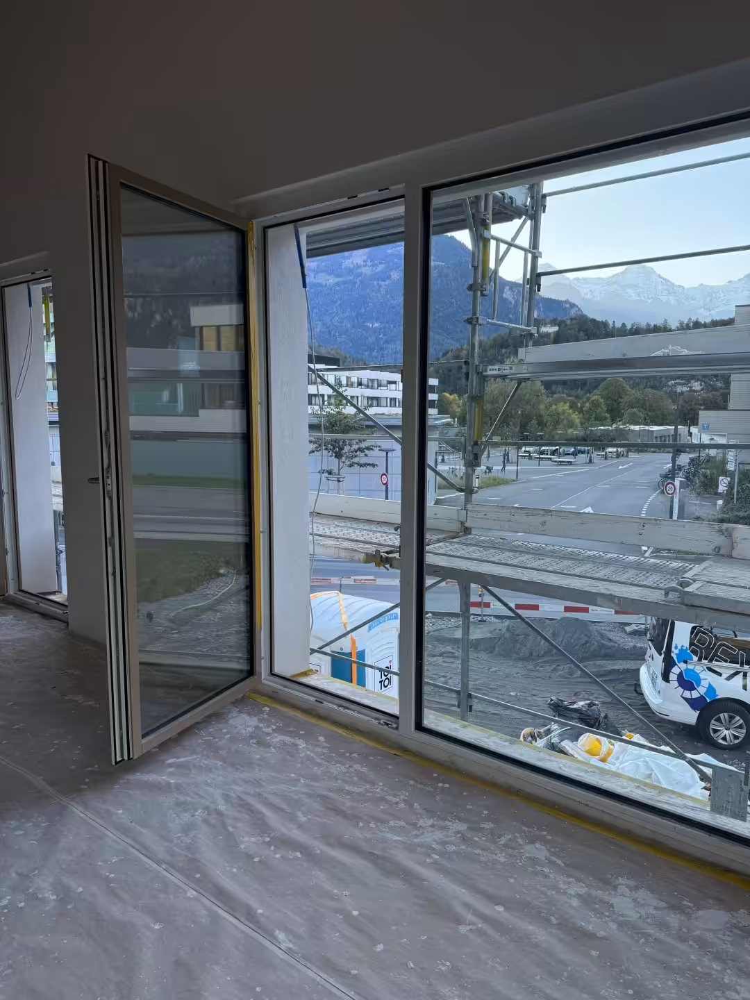 Blick aus einem Zimmer mit großen Fenstern auf eine Baustelle und Berge im Hintergrund.